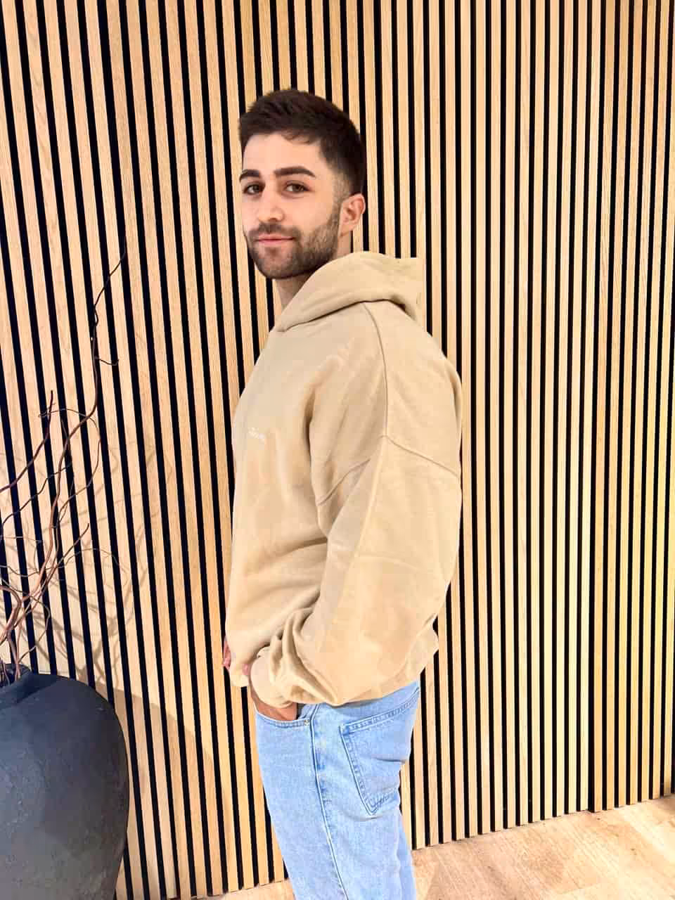 Oversize Hoodie - "Invictus" 5 Modisch gekleideter junger Mann vor Holzverkleidung, casual Streetwear, moderne Mode, urban Style.