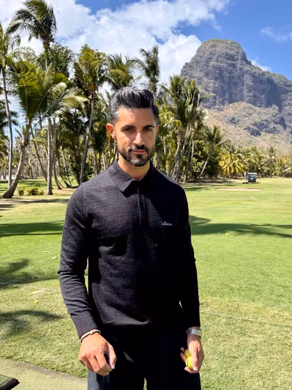 Mann im schwarzen Pullover im Garten mit Palmen und Berg im Hintergrund.