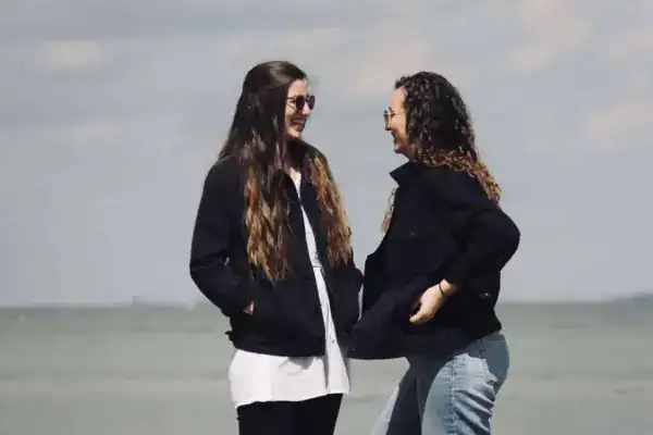 Freundliche Frauen lachen am Strand, Sommer, Mode, Freundschaft, sonniges Wetter, relaxte Atmosphäre.