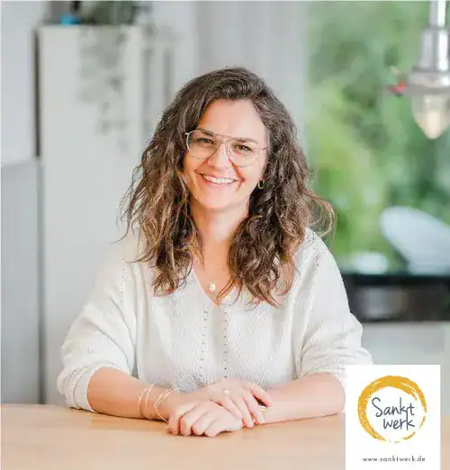 Authentische Frau mit Brille und freundlichem Lächeln, sitzend am Tisch in moderner Küche.