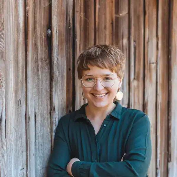 Hübsche junge Frau mit Brille vor Holzwand, freundliches Lächeln, natürliche Atmosphäre.