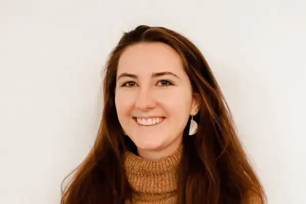 Hübsche Frau mit braunen Haaren und braunem Rollkragenpullover, lächelnd vor weißem Hintergrund.