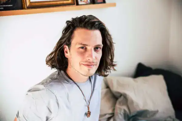 Relaxed junger mann sitzt auf einem sofa, hippie stil, langes haar, gemütliches wohnzimmer, freundliche ausstrahlung.