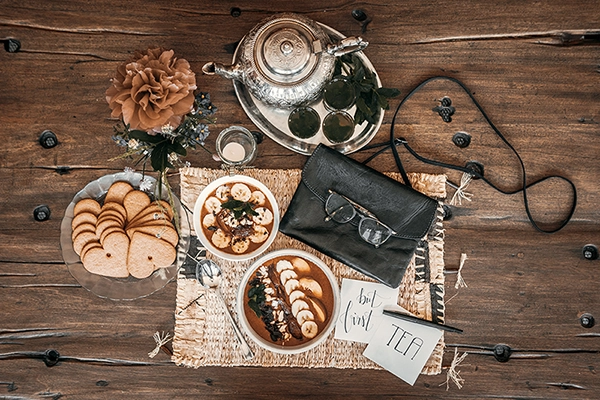 Draufsicht auf Holztisch bedeckt mit Müsli, Tee, schwarzer Tasche, Brille und mehr