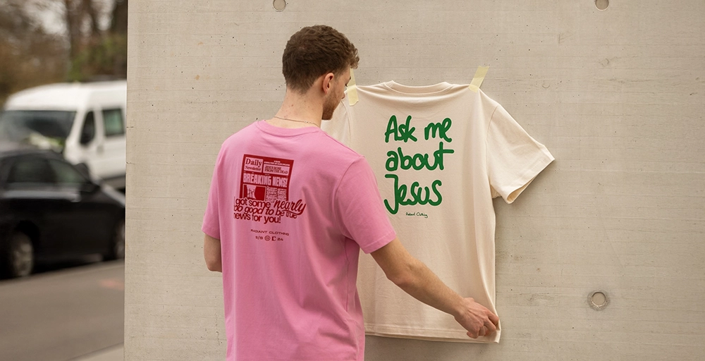 Mann mit pinkem Shirt klebt beiges ‚Ask me about Jesus‘ – Shirt an eine Betonwand