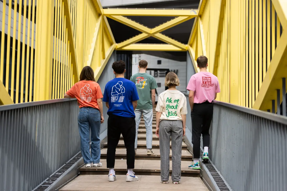 Fünf junge Erwachsene tragen verschieden bunte christliche Shirts und zeigen die Rückenseite