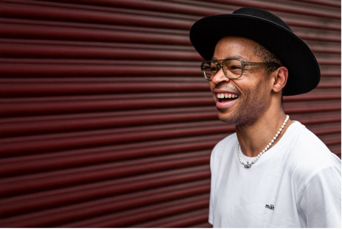 Model mit Hut steht lachend in weißem christlichen Shirt vor einer Garage