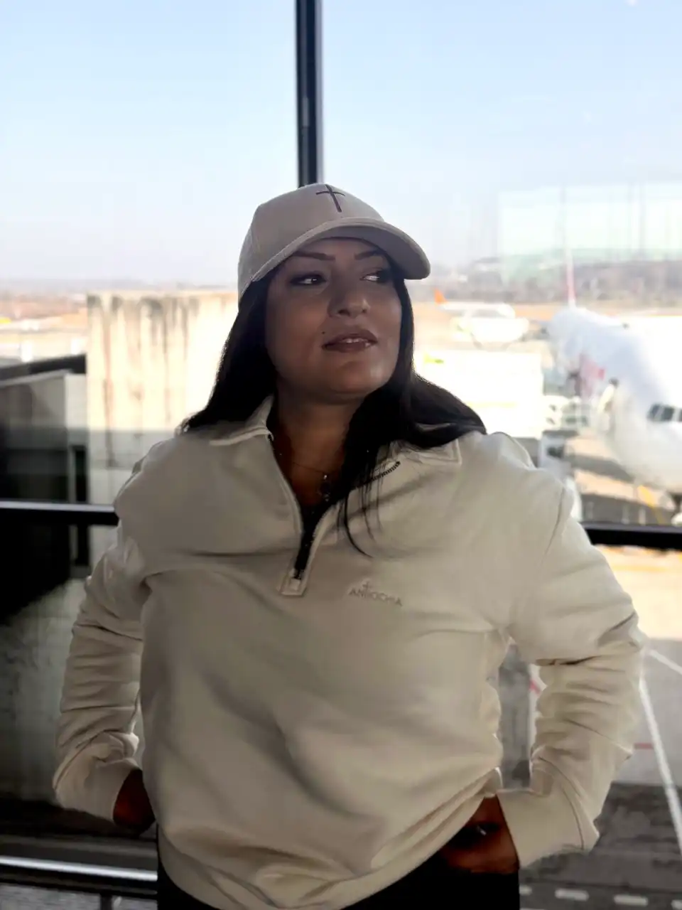 Kord Basecap - "Cross" 6 Gebündelte Frau in Kapuzenjacke und Baseballcap am Flughafen, Flugzeuge im Hintergrund.