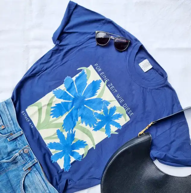 Ein blauer T-Shirt mit Blumenprint und Bibelvers, Sonnenbrille und Tasche auf weißem Stoff.