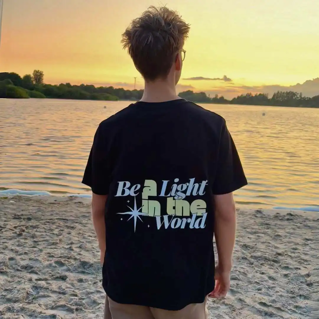 BE A LIGHT – Christliches T-Shirt 1 Der junge Mann trägt ein schwarzes T-Shirt mit dem Aufdruck "Be a Light in the World" am Fluss bei S.