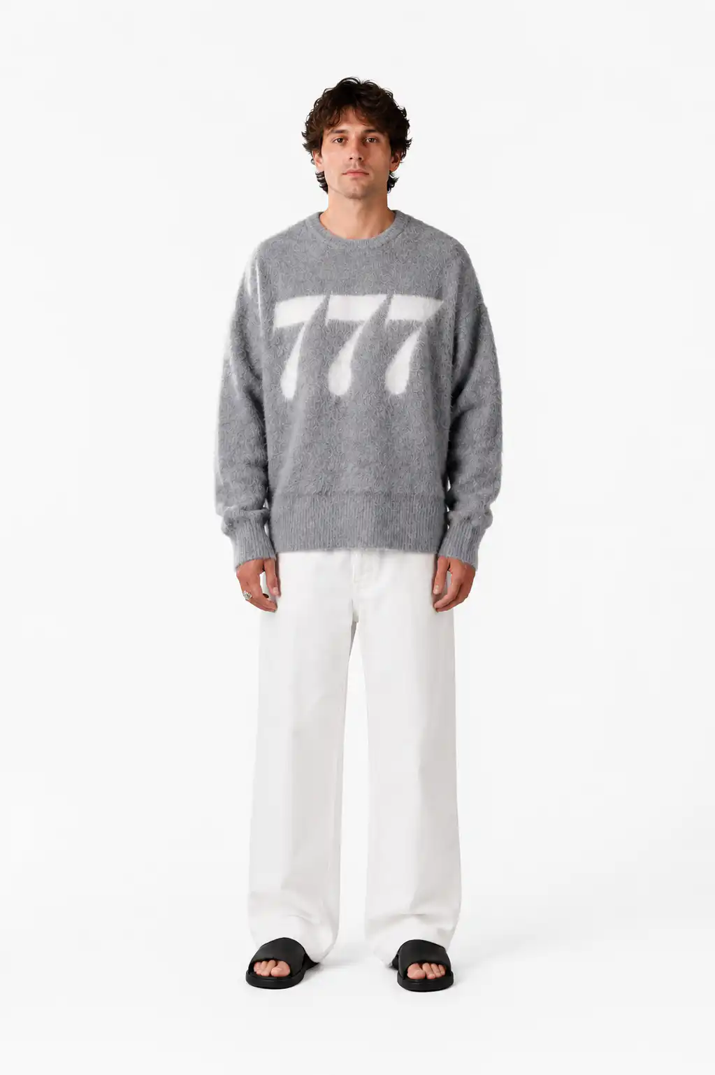 Der Mann trägt einen grauen Pullover mit weißen 777-Print, weiße Hosen und schwarze Sandalen.