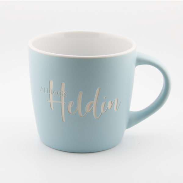 Hellblaue Tasse mit goldener Beschriftung 'Alltags Heldin' auf weißem Hintergrund.