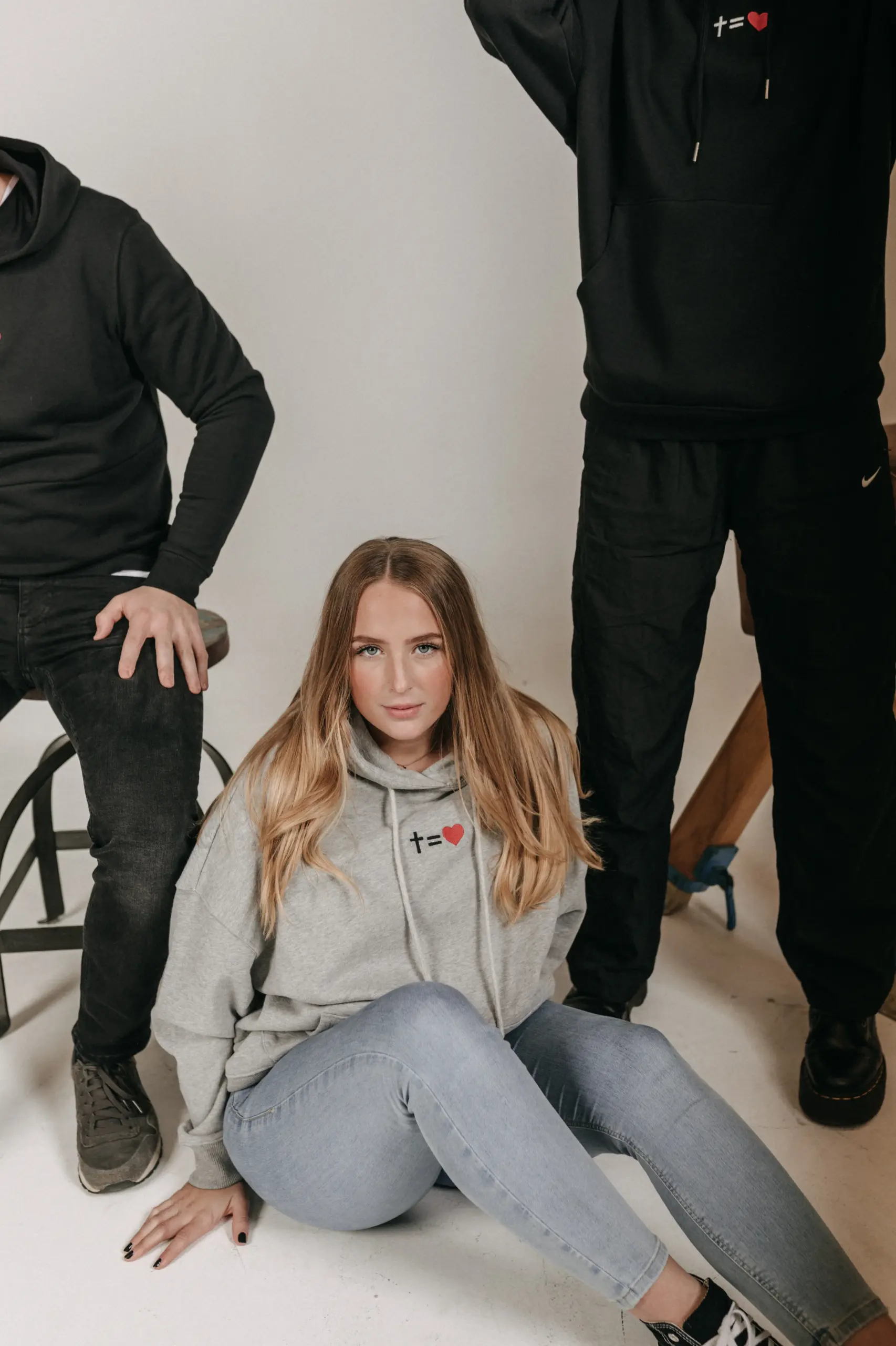 kreuzgleichliebe Hoodie Style Gratitude - unisex 18 Junge Frau in grauem Hoodie sitzt auf Boden, umgeben von zwei Männern in schwarzen Hoodies.