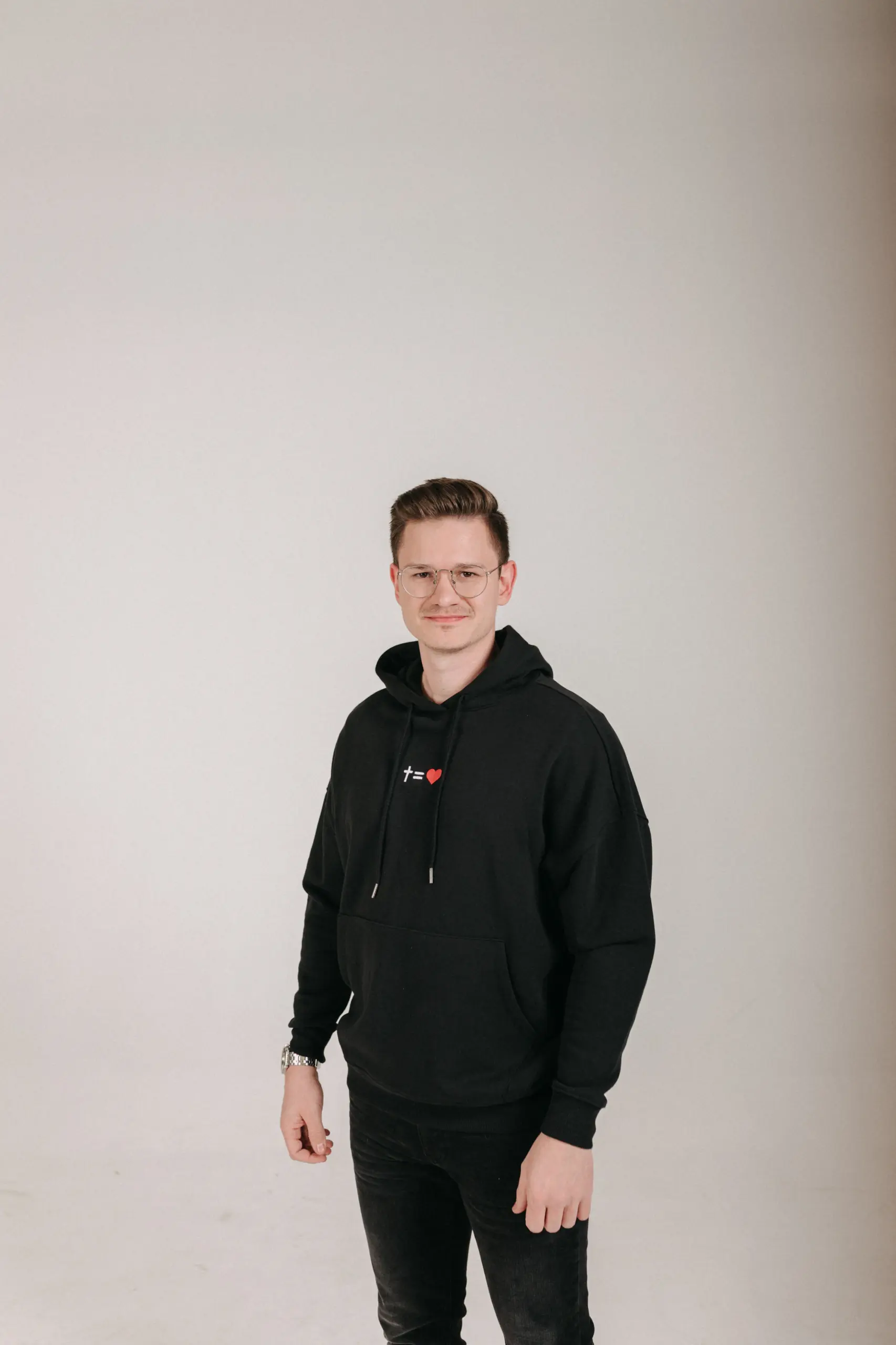 kreuzgleichliebe Hoodie Style Gratitude - unisex 17 Junger Mann trägt schwarze Kapuzenjacke mit christlichem Logo, lächelnd, neutraler Hintergrund.
